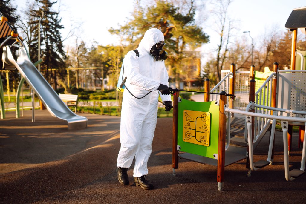 Man Wearing Protective Suit Disinfecting The Playground In The Sun With Spray. Covid 19. Cleaning.