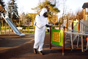 Man Wearing Protective Suit Disinfecting The Playground In The Sun With Spray. Covid 19. Cleaning.