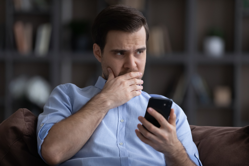 Man In A Blue Shirt Sitting On A Couch, Looking At His Smartphone With A Concerned Expression, Partially Covering His Mouth With His Hand. Shelves With Books And Decor Are In The Background.